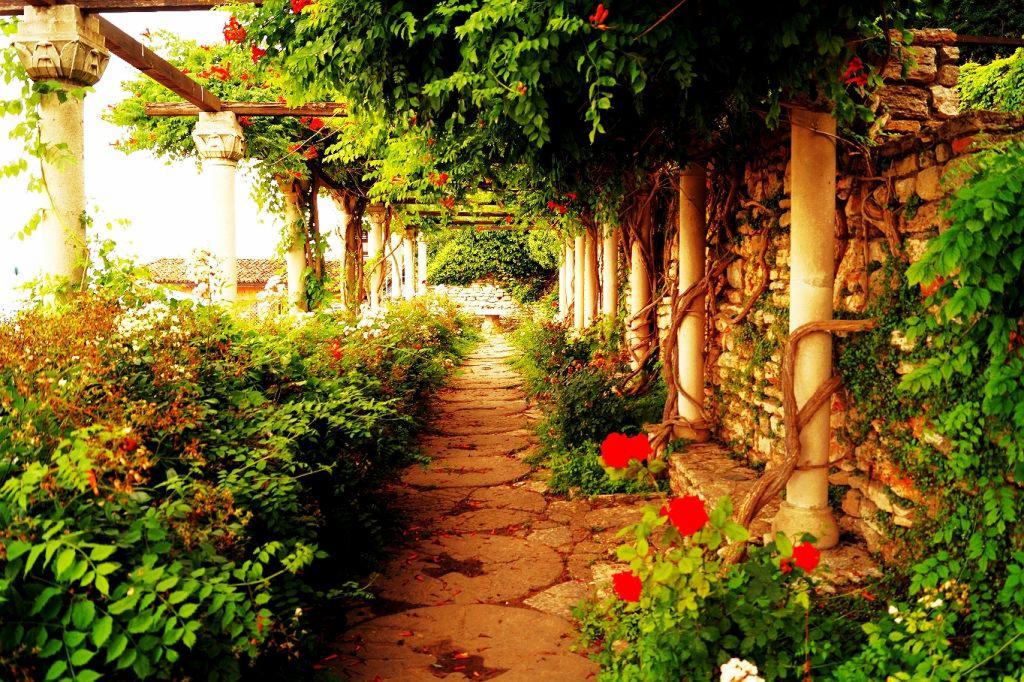 Urokliwa aleja w ogrodzie z kamienną ścieżką, kolumnami oplecionymi pnączami i kwitnącymi roślinami, tworząca malowniczy tunel roślinny.