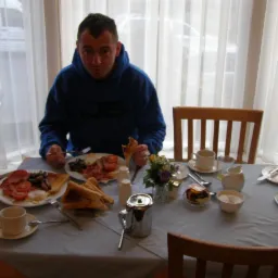 Mężczyzna w niebieskiej bluzie siedzi przy stole zastawionym śniadaniem: jajka sadzone z boczkiem i pomidorami, tosty, kawa, deser; za nim okno z białymi zasłonami.