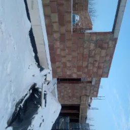 Wznoszenie murów domu z czerwonej cegły w zimowej scenerii, widoczne palety cegieł pod śniegiem i zbrojenie.