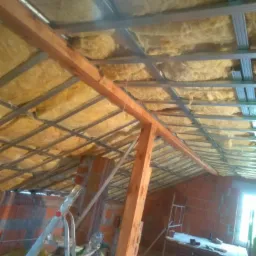 Poddasze w trakcie ocieplania wełną mineralną, widoczne metalowe profile konstrukcyjne i fragment ściany z cegły.