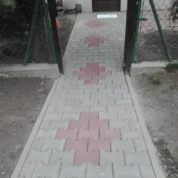 Ścieżka z szarej kostki brukowej z czerwonymi akcentami prowadząca do brązowych drzwi, ogrodzona siatką z tabliczką 'Uwaga Pies'.