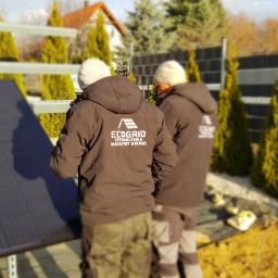 ECOGRID Bartosz Choromański - Dwaj instalatorzy w kurtkach z logo firmy 'Ecogrid' montują panele słoneczne na metalowej konstrukcji w ogrodzie z tujami i domem w tle.
