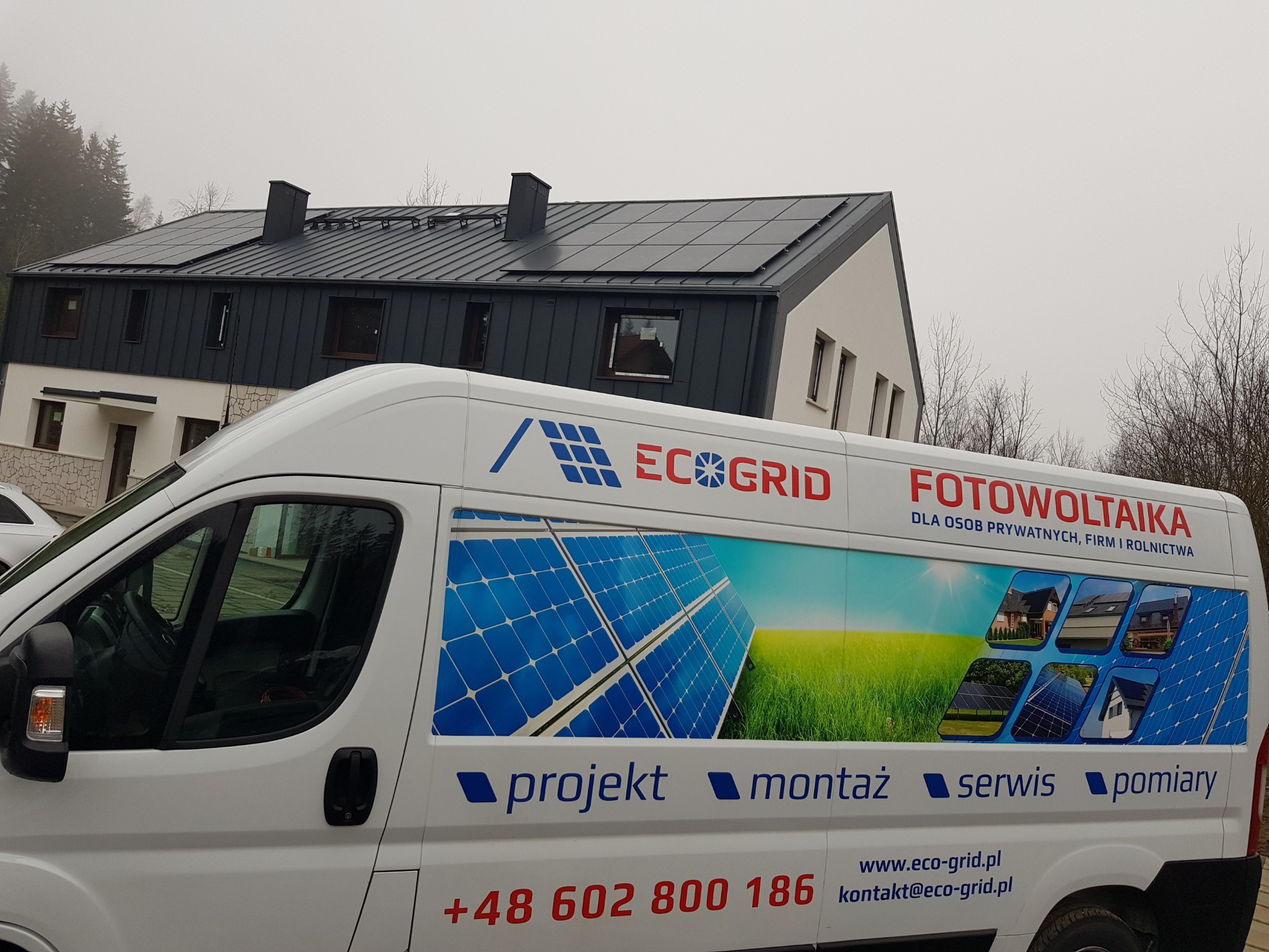 Biały samochód dostawczy z reklamą firmy Eco Grid i hasłem 'Fotowoltaika dla osób prywatnych, firm i rolnictwa' zaparkowany przed domem z panelami słonecznymi na dachu.