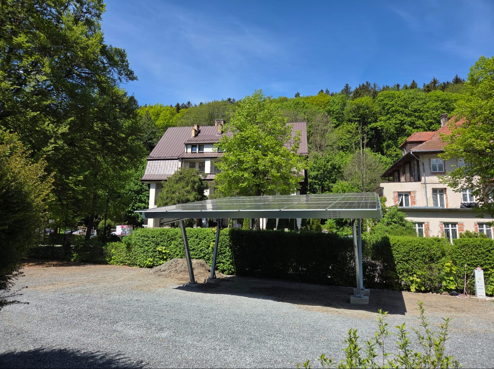 Carport z panelami fotowoltaicznymi na metalowej konstrukcji, zadaszający żwirowy parking, otoczony bujną zielenią żywopłotów i drzew, z widocznymi budynkami w tle, w słoneczny dzień.