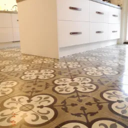 Wzorzyste, beżowo-białe płytki podłogowe w kuchni, widok z niskiego kąta, z fragmentem białej wyspy kuchennej z szarym blatem i brązowymi uchwytami.