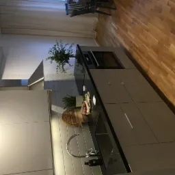 Nowoczesna kuchnia z szarymi szafkami, czarnym blatem, białymi płytkami na ścianie i drewnianą podłogą. Na blacie widoczne zioła w doniczkach, deska do krojenia i dekoracyjne gałązki.