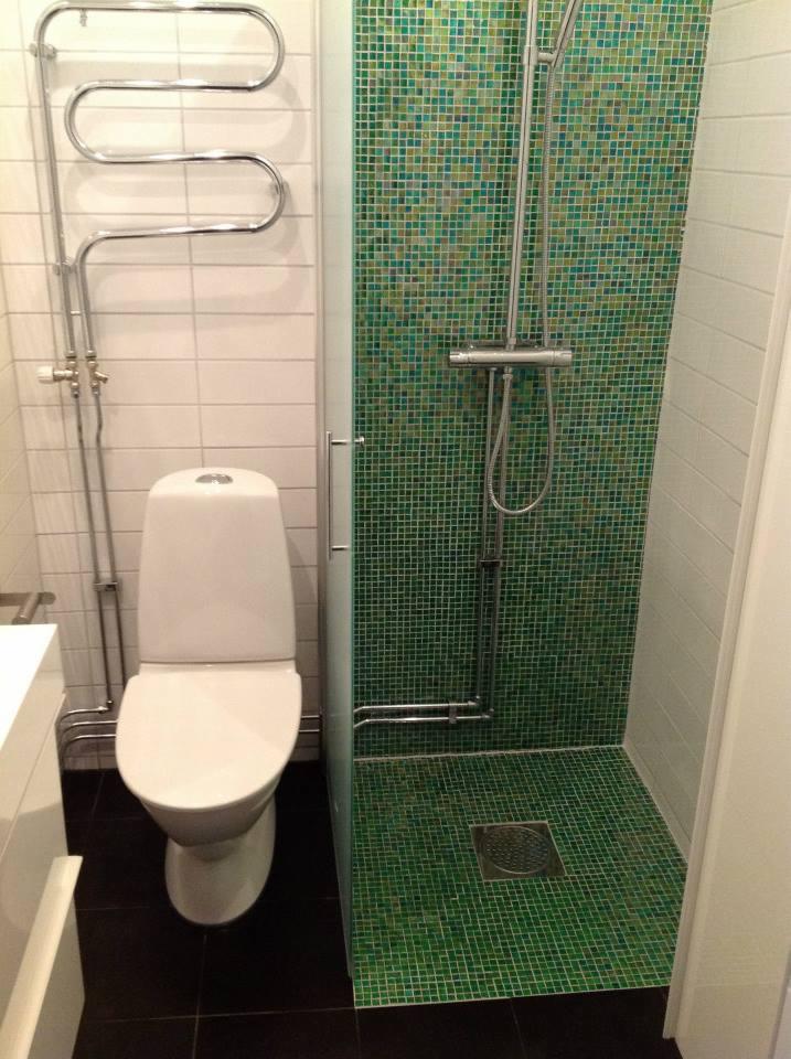 Mała łazienka z kabiną prysznicową wyłożoną zielono-niebieską mozaiką, białą toaletą i grzejnikiem drabinkowym na ścianie z białych płytek.