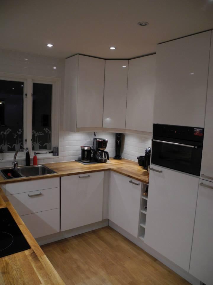 Jasna kuchnia z białymi, błyszczącymi szafkami, drewnianym blatem, białymi płytkami ściennymi i drewnianą podłogą; widoczne ekspresy do kawy i piekarnik.