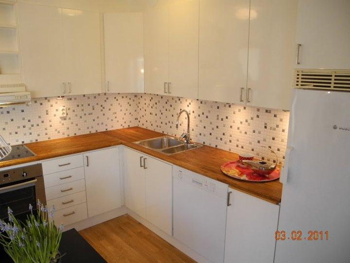 Jasna kuchnia w kształcie litery L z drewnianym blatem, mozaiką na ścianie i białymi frontami szafek, widoczny zlew, bateria i płyta grzewcza.