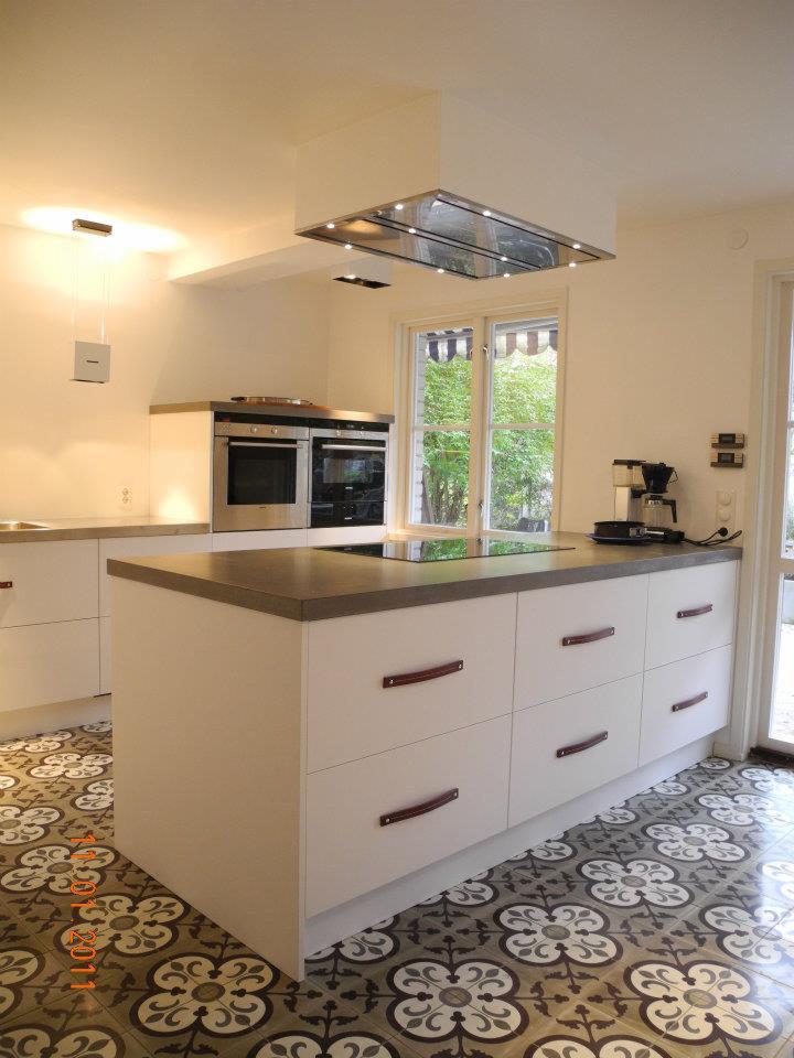 Jasna kuchnia z wyspą, szary blat, białe szafki z brązowymi uchwytami, wzorzysta podłoga, okap nad płytą grzewczą, ekspres do kawy na blacie.