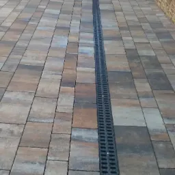 Ułożona kostka brukowa w odcieniach brązu i szarości z centralnie umieszczonym, czarnym odpływem liniowym.