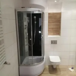 Nowoczesna łazienka z narożną kabiną prysznicową z hydromasażem, białą ceramiką i drewnopodobną podłogą, oświetlona wiszącą żarówką.