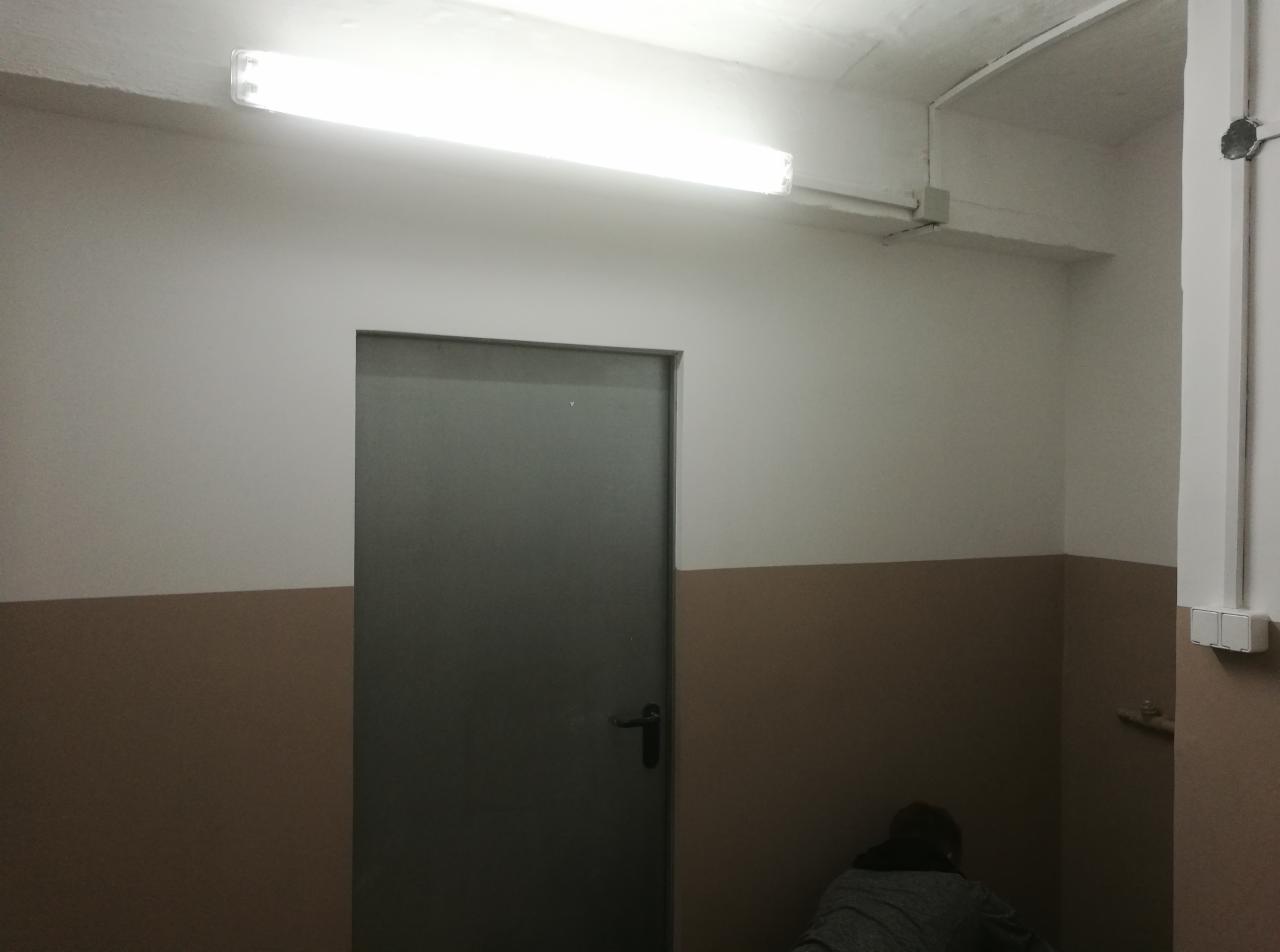 Korytarz w trakcie prac remontowych: świeżo pomalowane ściany do połowy na biało, poniżej brązowy pas, widoczna szara metalowa futryna drzwi i wystająca lampa jarzeniowa pod sufitem, osoba w rogu...