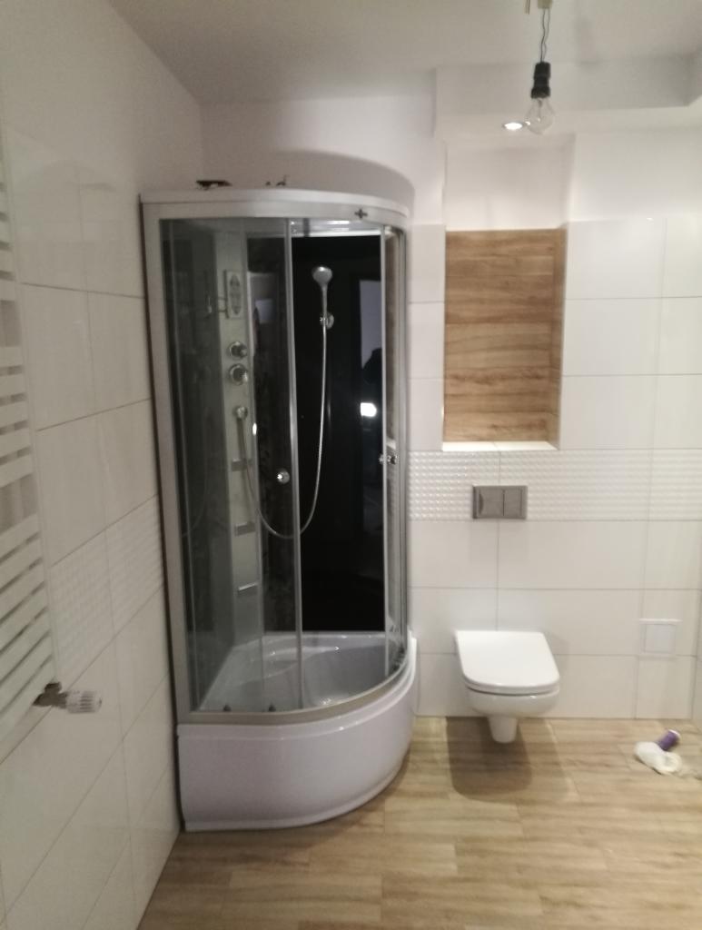 Nowoczesna łazienka z narożną kabiną prysznicową z hydromasażem, białą ceramiką i drewnopodobną podłogą, oświetlona wiszącą żarówką.