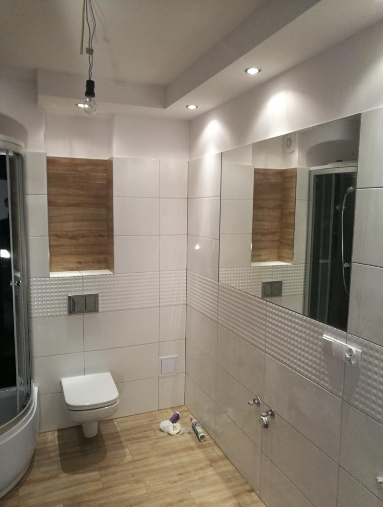 Wykończona łazienka z białymi płytkami, drewnopodobną podłogą i wnęką z drewnianą okładziną. Widoczna toaleta, kabina prysznicowa i duże lustro. Na podłodze leżą narzędzia i materiały budowlane.