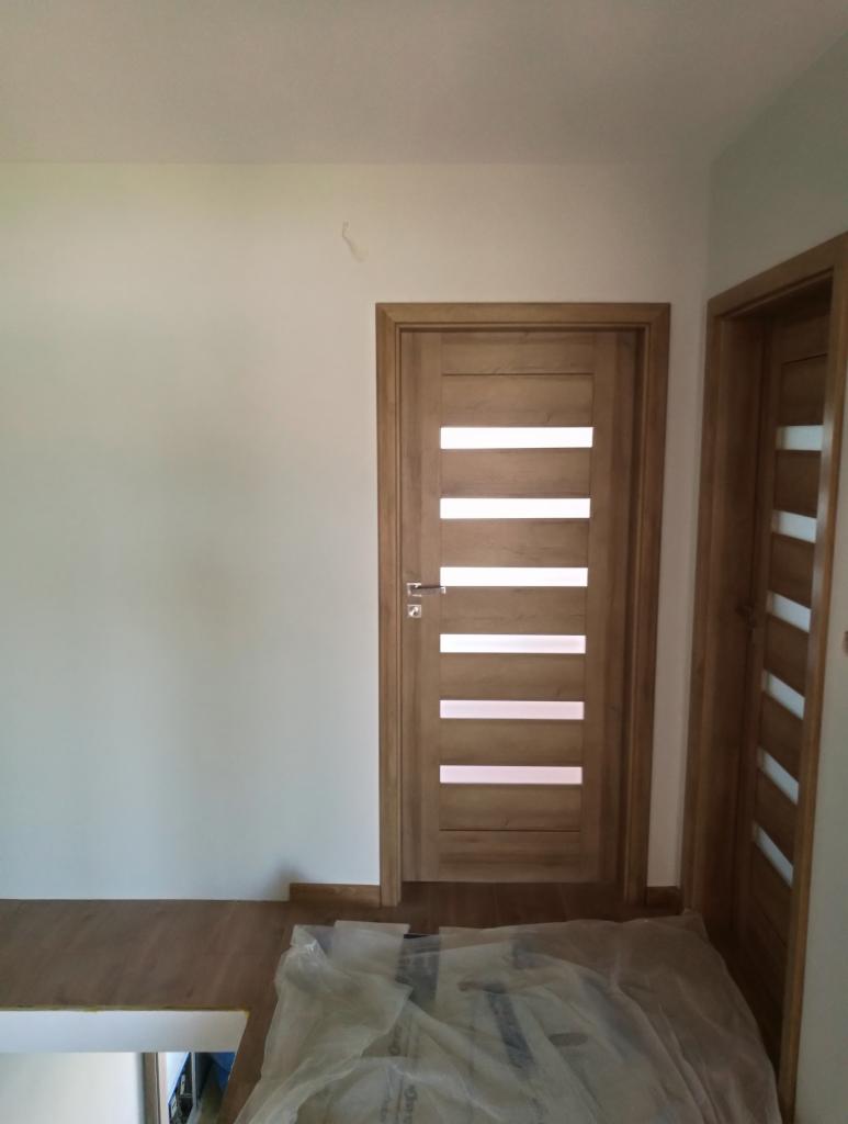 Zamontowane drewniane drzwi wewnętrzne z poziomymi przeszkleniami, widoczne zabezpieczenie podłogi folią malarską, ściana pomalowana na biało.