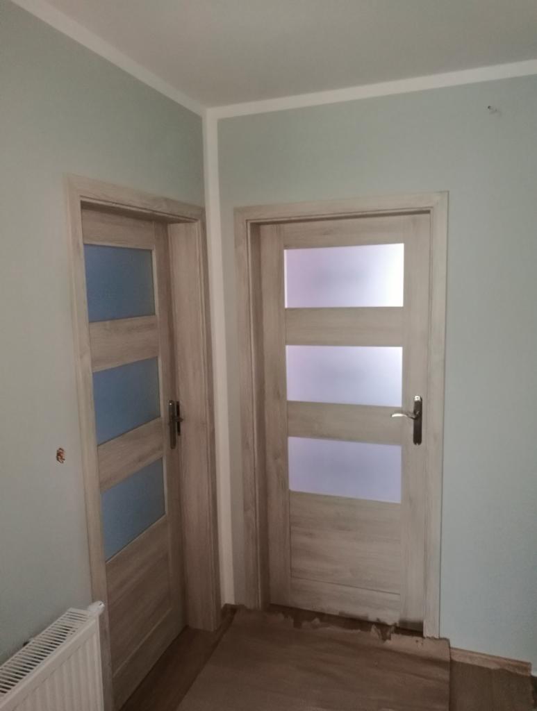 Dwa drewnopodobne drzwi wewnętrzne z trzema poziomymi szybami z mlecznego szkła, zamontowane w ścianie o kolorze jasnej zieleni, widoczne fragmenty podłogi i grzejnika.