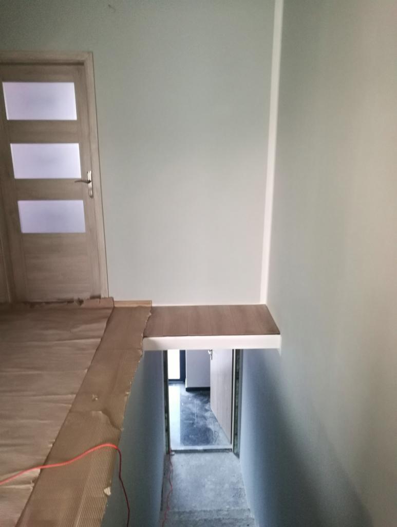 Przygotowanie do remontu: zabezpieczona podłoga kartonem, widok na schody w dół z zamontowaną półką nad wejściem, świeżo pomalowane ściany na jasny kolor.