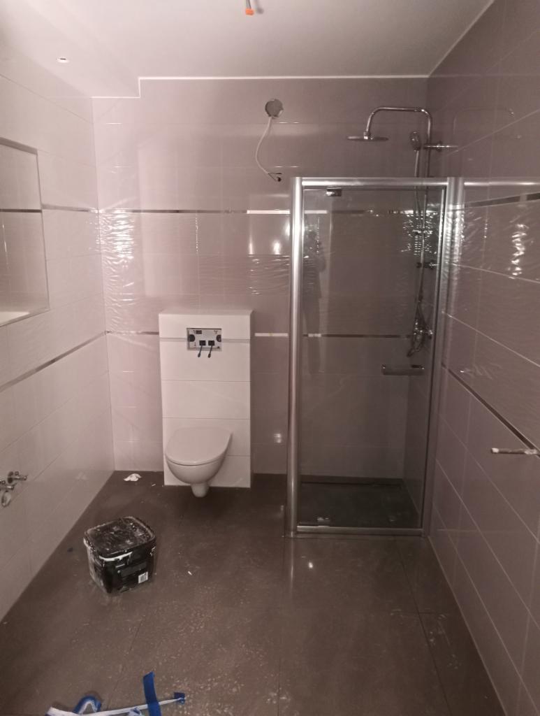 Niedokończona łazienka z szarymi płytkami, kabiną prysznicową i podwieszanym WC. Widoczny pojemnik z farbą i narzędzia na podłodze.