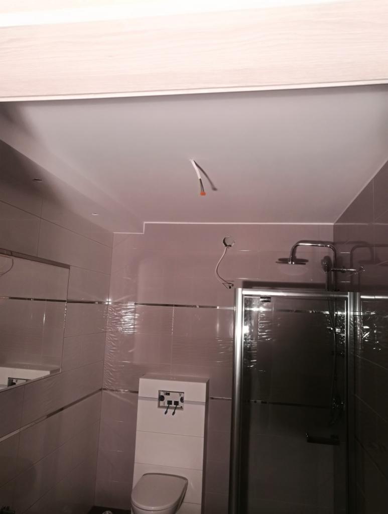 Łazienka w trakcie remontu: widoczny biały sufit napinany z wystającymi kablami, częściowo wykończone ściany z jasnymi płytkami oraz kabina prysznicowa z metalową ramą.