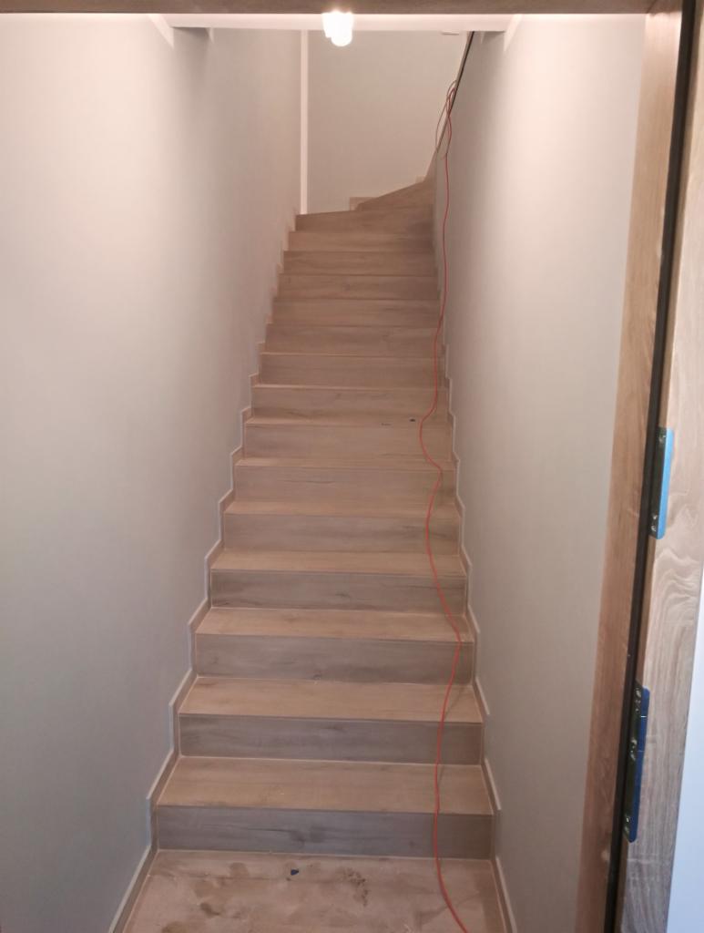 Schody wyłożone panelami imitującymi drewno, prowadzące w górę, z widocznym czerwonym kablem biegnącym wzdłuż ściany i fragmentem ościeżnicy drzwi po prawej stronie.