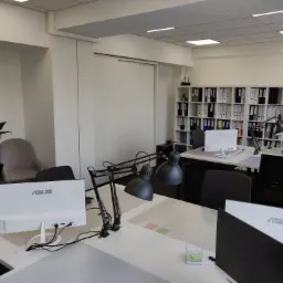 Jasne biuro z białymi biurkami, lampami biurkowymi i monitorami ASUS, regał z segregatorami w tle, minimalistyczny wystrój.