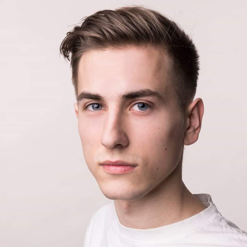 Portret młodego mężczyzny z krótkimi brązowymi włosami zaczesanymi na bok i jasnymi oczami, na jasnym tle.