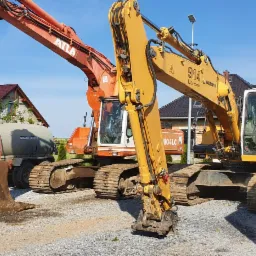 Dwie koparki, pomarańczowa Atlas i żółta O&K, stoją na żwirowym placu; w tle domy i przyczepa z zielonym zbiornikiem.