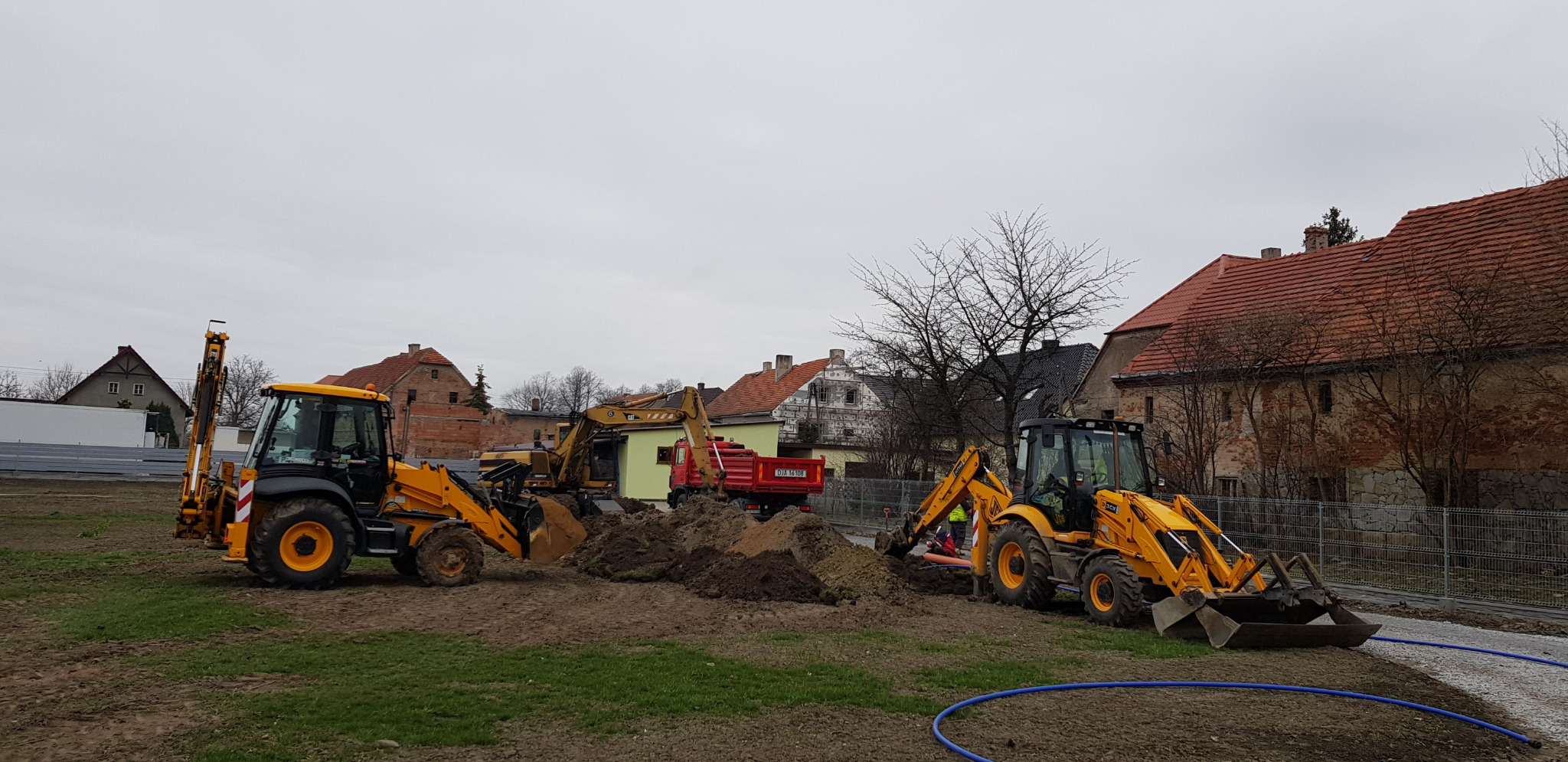Dwie żółte koparko-ładowarki JCB pracują na placu budowy; w tle widoczny czerwony wywrotka, budynki mieszkalne i pochmurne niebo.