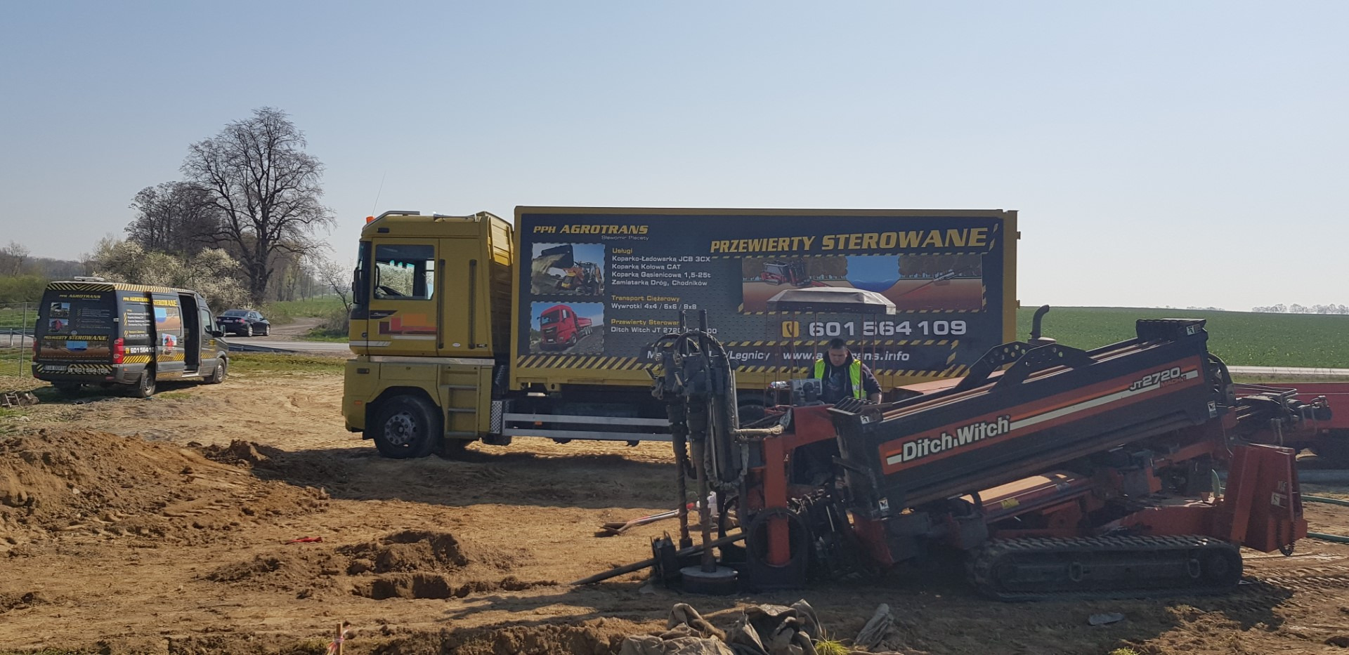 Żółta ciężarówka z reklamą firmy PPH Agrotrans i sprzęt Ditch Witch JT2720 do przewiertów sterowanych na placu budowy, widoczny pracownik w kamizelce odblaskowej, w tle samochód dostawczy tej samej...