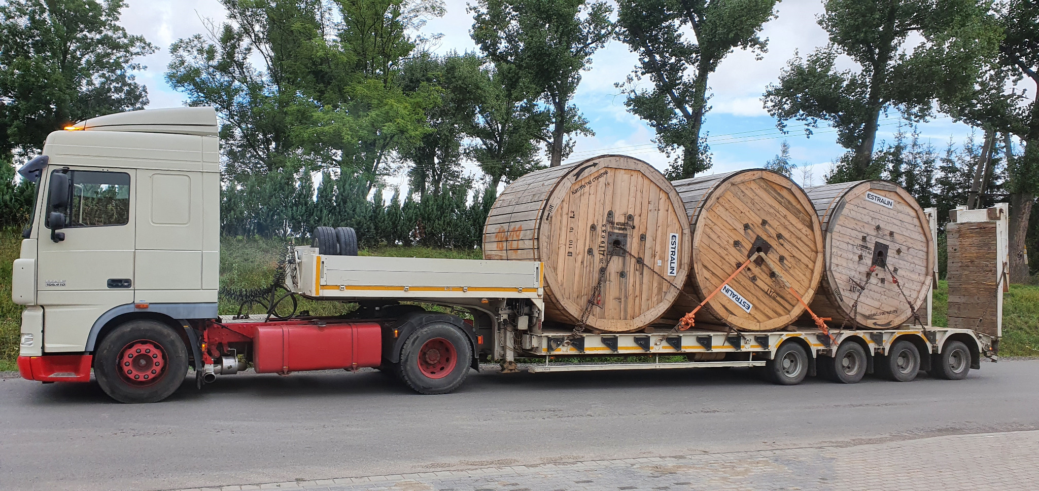 Ciężarówka z naczepą przewozi trzy duże, drewniane szpule z nawiniętym kablem na tle drzew i pochmurnego nieba.