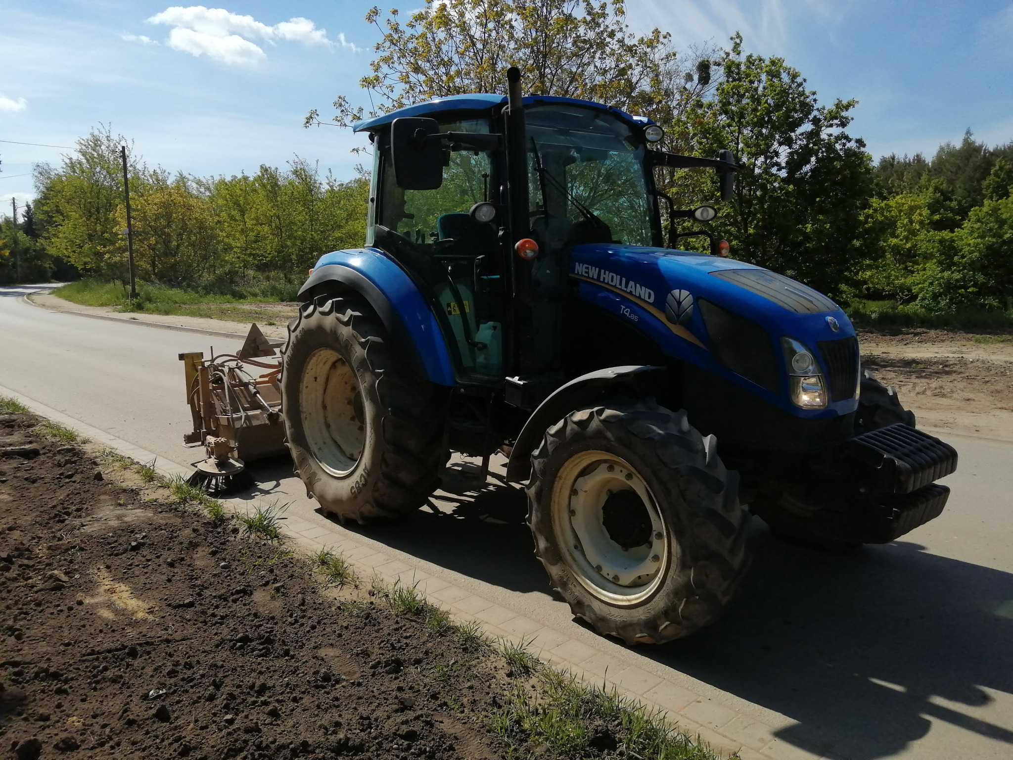Niebieski traktor New Holland T4.as z zamontowanym sprzętem roboczym przy krawędzi asfaltowej drogi, obok świeżo przygotowany pas ziemi.