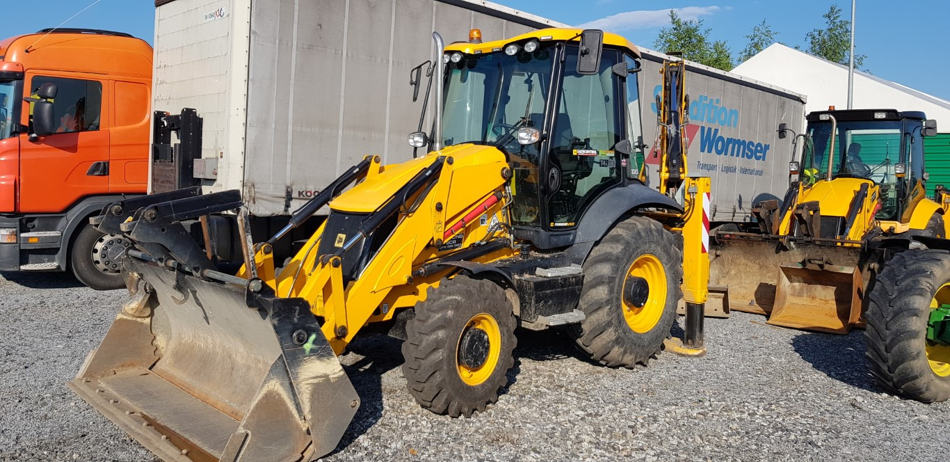 Żółta koparko-ładowarka JCB na żwirowym placu, obok pomarańczowej ciężarówki Scania i naczepy Wormser, z drugą koparko-ładowarką w tle.