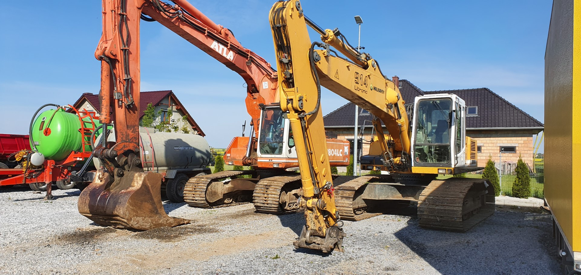 Dwie koparki, pomarańczowa Atlas i żółta O&K, stoją na żwirowym placu; w tle domy i przyczepa z zielonym zbiornikiem.