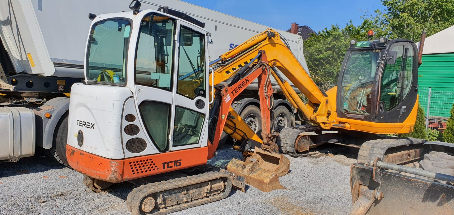 Dwa minikoparki: biała Terex TC16 i żółta, stoją na szarym żwirze obok ciężarówki z naczepą. Pierwsza minikoparka ma podniesione ramię kopiące, a druga stoi z opuszczonym ramieniem.