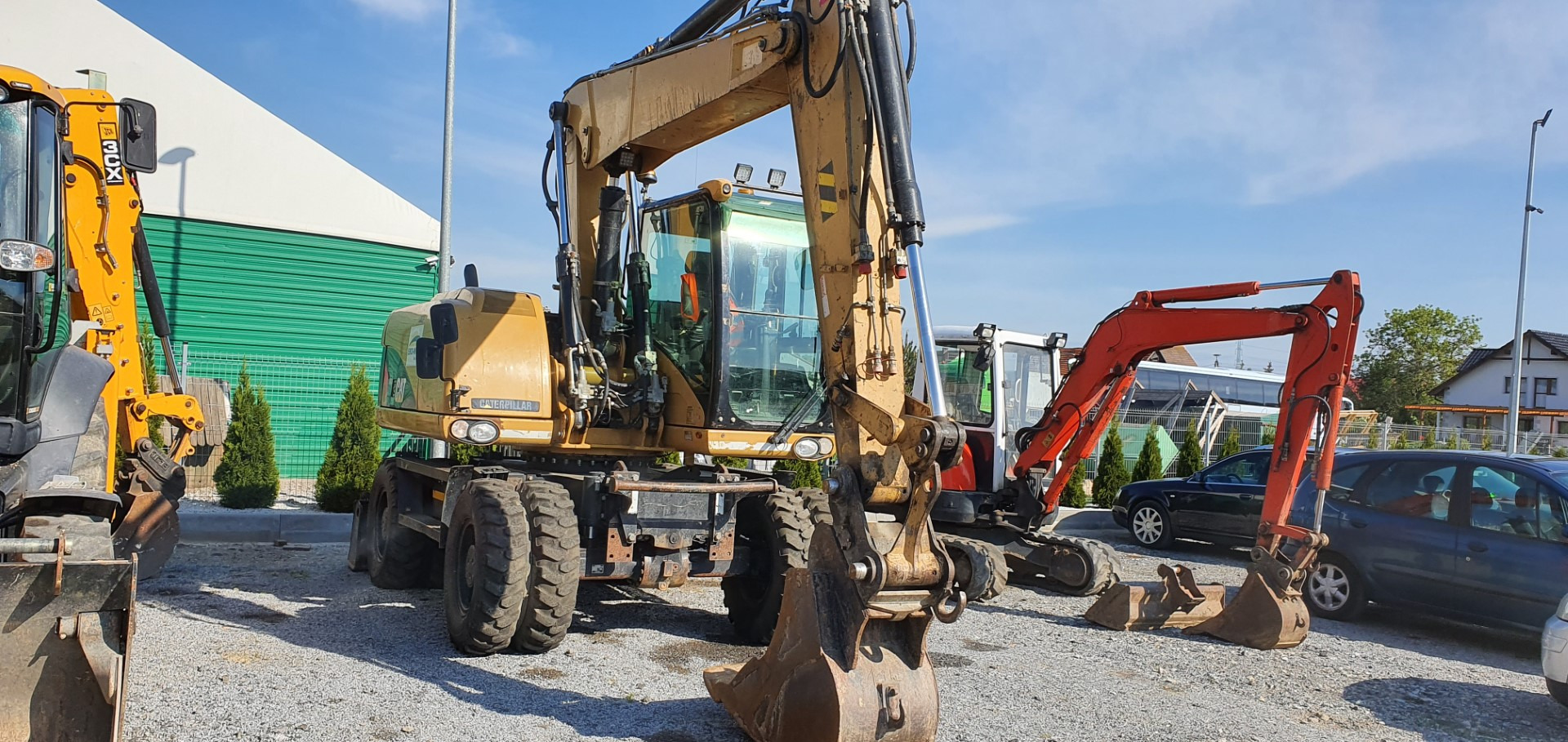 Żółta koparka kołowa Caterpillar na żwirowym placu, obok zaparkowane inne maszyny budowlane i samochody, w tle zielony budynek.