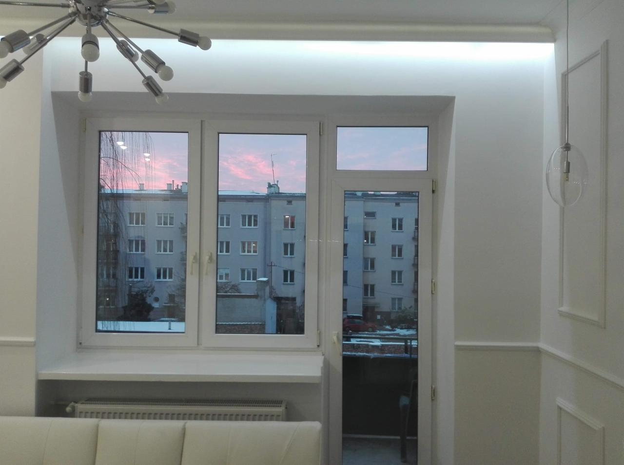 Nowoczesne, białe wnętrze z trzyskrzydłowym oknem z widokiem na budynki o wschodzie słońca, dekoracyjna lampa sufitowa i wisząca lampa kulista.