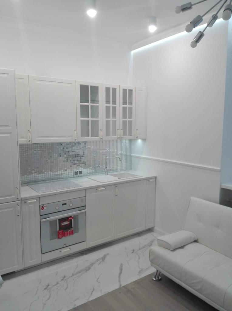 Jasna kuchnia z białymi szafkami, mozaiką nad blatem i marmurową podłogą, uzupełniona białą sofą.