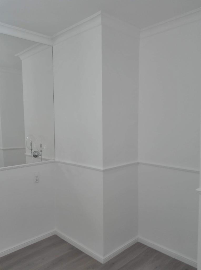 Białe ściany z dekoracyjnymi listwami przypodłogowymi i sufitowymi, lustro z kinkietem w rogu pokoju, szare panele podłogowe.