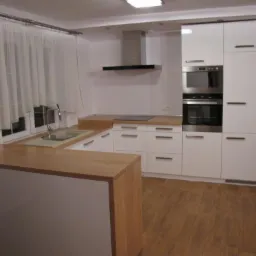 Jasna kuchnia z białymi szafkami, drewnianym blatem i oknem z firankami, widok na zabudowę kuchenną z wyspą.