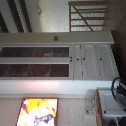 Biała zabudowa meblowa z przeszklonymi i pełnymi szafkami, zamontowana pod sufitem w salonie z telewizorem i drewnianymi schodami w tle.