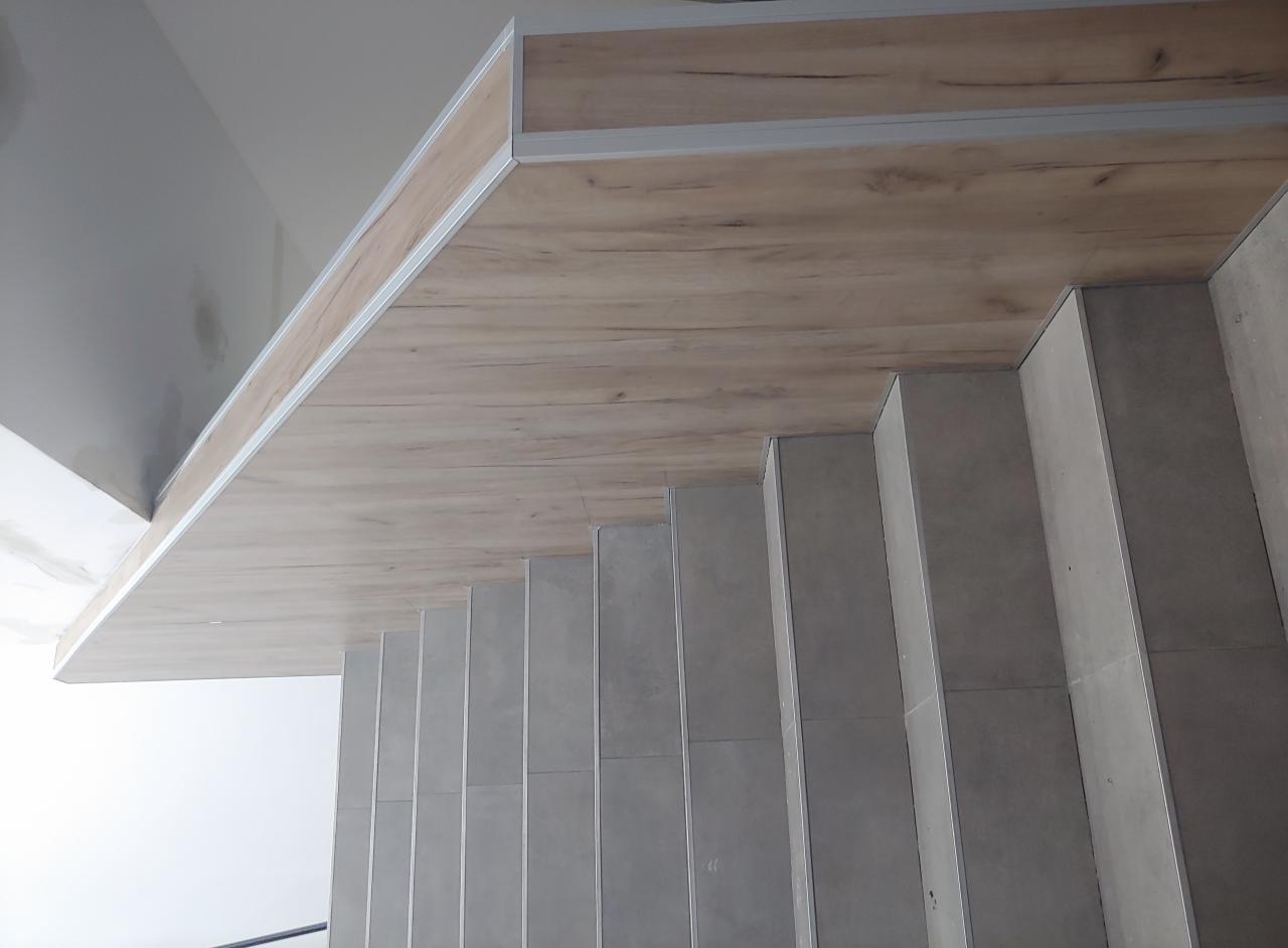 Wykończone schody z szarych płytek, drewnopodobna okładzina sufitu i srebrne listwy wykończeniowe.