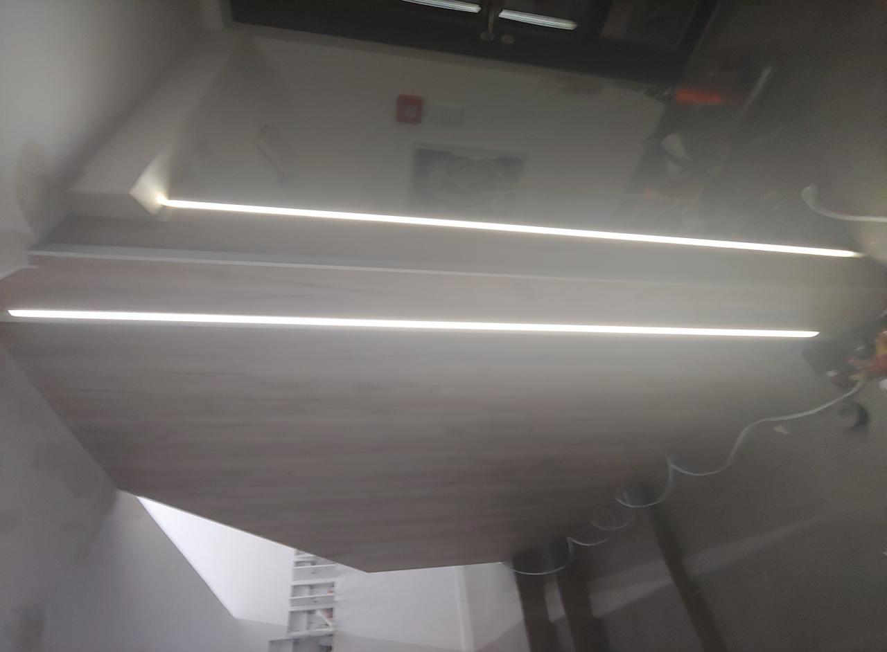 Fragment sufitu z dwiema liniami równoległego oświetlenia LED, widoczne kable, szara ściana i białe elementy konstrukcyjne w tle.