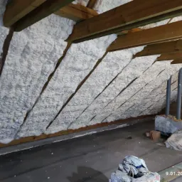 Poddasze ocieplone pianką PUR, widoczne drewniane belki konstrukcyjne, szara podłoga, fragment muru z rurami, data 9.01.2023 w rogu.