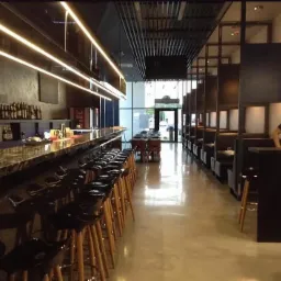 Wnętrze lokalu usługowego z długim barem z marmurowym blatem i rzędem czarnych hokerów na drewnianych nogach, oświetlone ledowymi listwami, w tle kabiny dla gości i obsługa w czarnej koszulce.