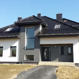 Nowoczesny dom jednorodzinny z szarą elewacją, białymi elementami i ciemną dachówką, widoczny z perspektywy ścieżki z kamyczków, na tle błękitnego nieba.