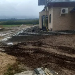 Budowa schodów zewnętrznych z kostki granitowej przy domu z jasnej cegły; widoczne narzędzia, piasek i błoto na placu budowy w pochmurny dzień.