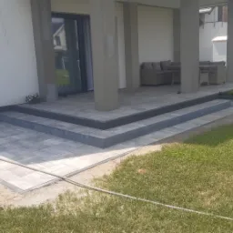 Nowoczesny taras z szarej kostki brukowej z trzema stopniami, prowadzący do zadaszonego patio z meblami ogrodowymi, widok z perspektywy trawnika.