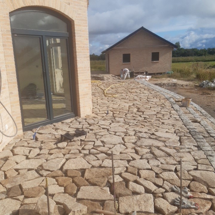 Nieregularna kamienna nawierzchnia tarasu w trakcie budowy, widoczne narzędzia, fragment budynku z cegły i pracownik w tle.