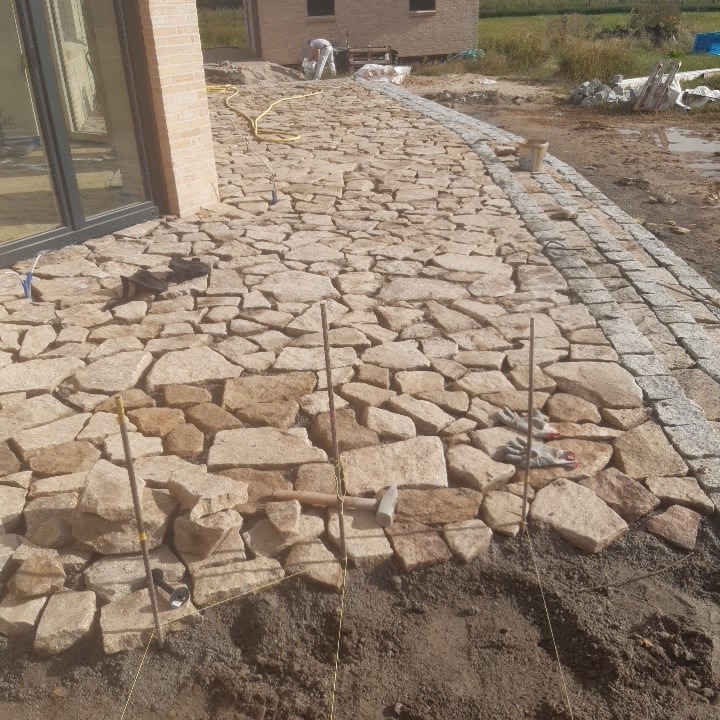 Układanie nieregularnej kostki kamiennej w odcieniach beżu i brązu na ścieżce ogrodowej, widoczne narzędzia pracy, w tle budynek w trakcie budowy.